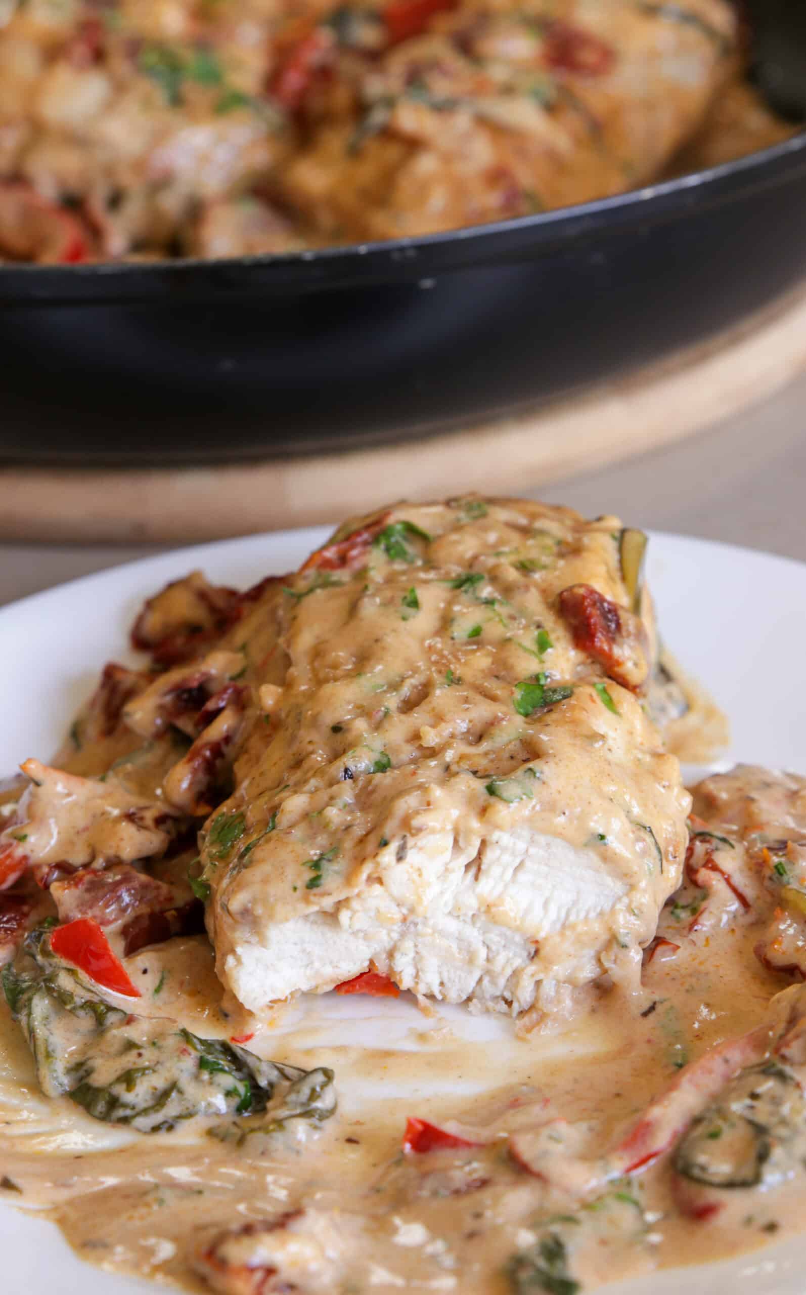 A plate of creamy Tuscan chicken: a sliced chicken breast covered in creamy sauce, garnished with chopped herbs, with spinach, sun-dried tomatoes, and bell peppers. A skillet with more chicken is in the background.