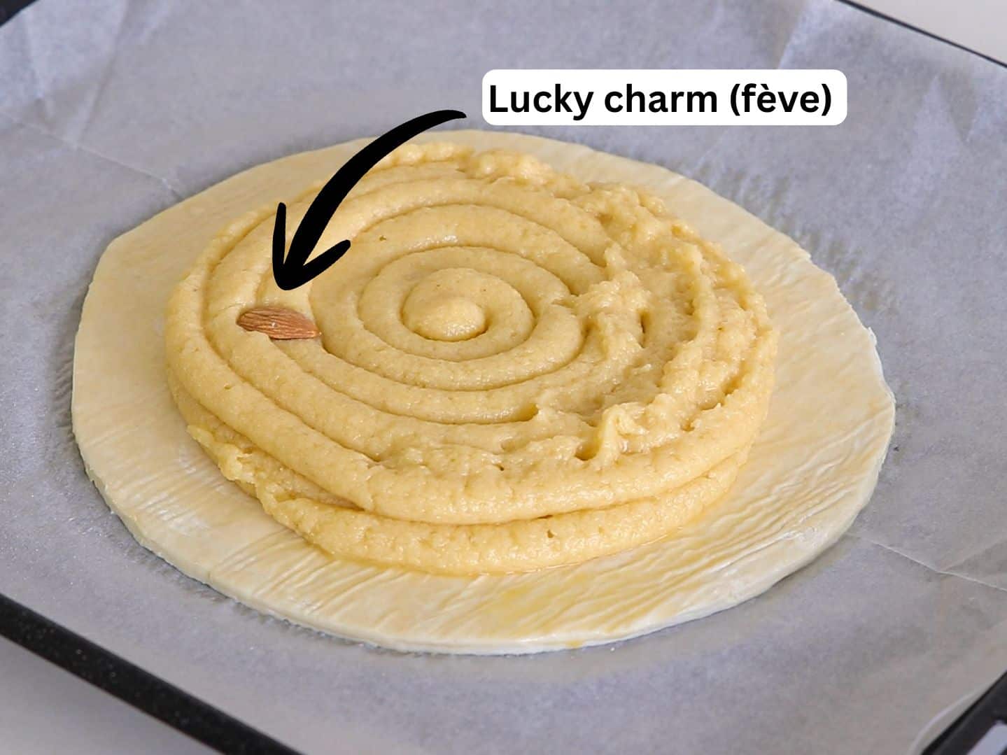 A round pastry with spiral filling sits on parchment paper. An almond, labeled “Lucky charm (fève)” with an arrow, is placed on top of the filling before baking.