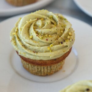 A pistachio cupcake topped with green pistachio frosting swirled in a rose shape and sprinkled with crushed pistachios, served on a white plate.