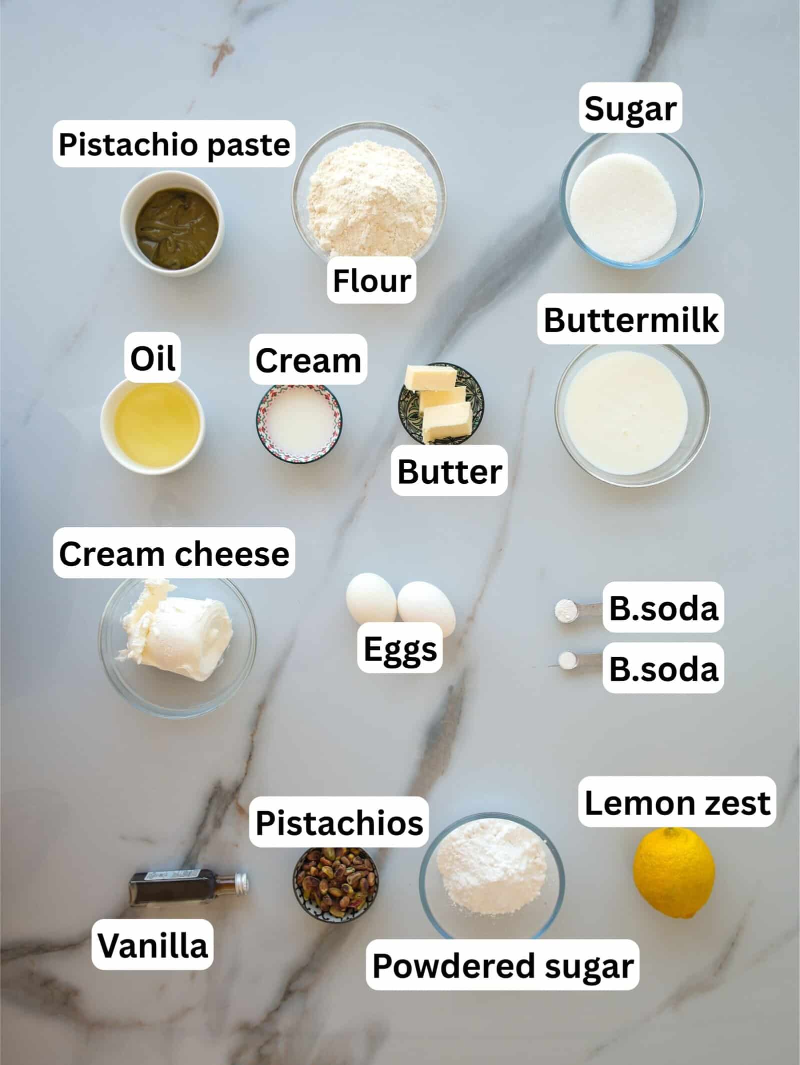 A marble countertop with labeled bowls containing pistachio paste, flour, sugar, buttermilk, oil, cream, butter, cream cheese, eggs, baking soda, lemon zest, vanilla, pistachios, and powdered sugar.