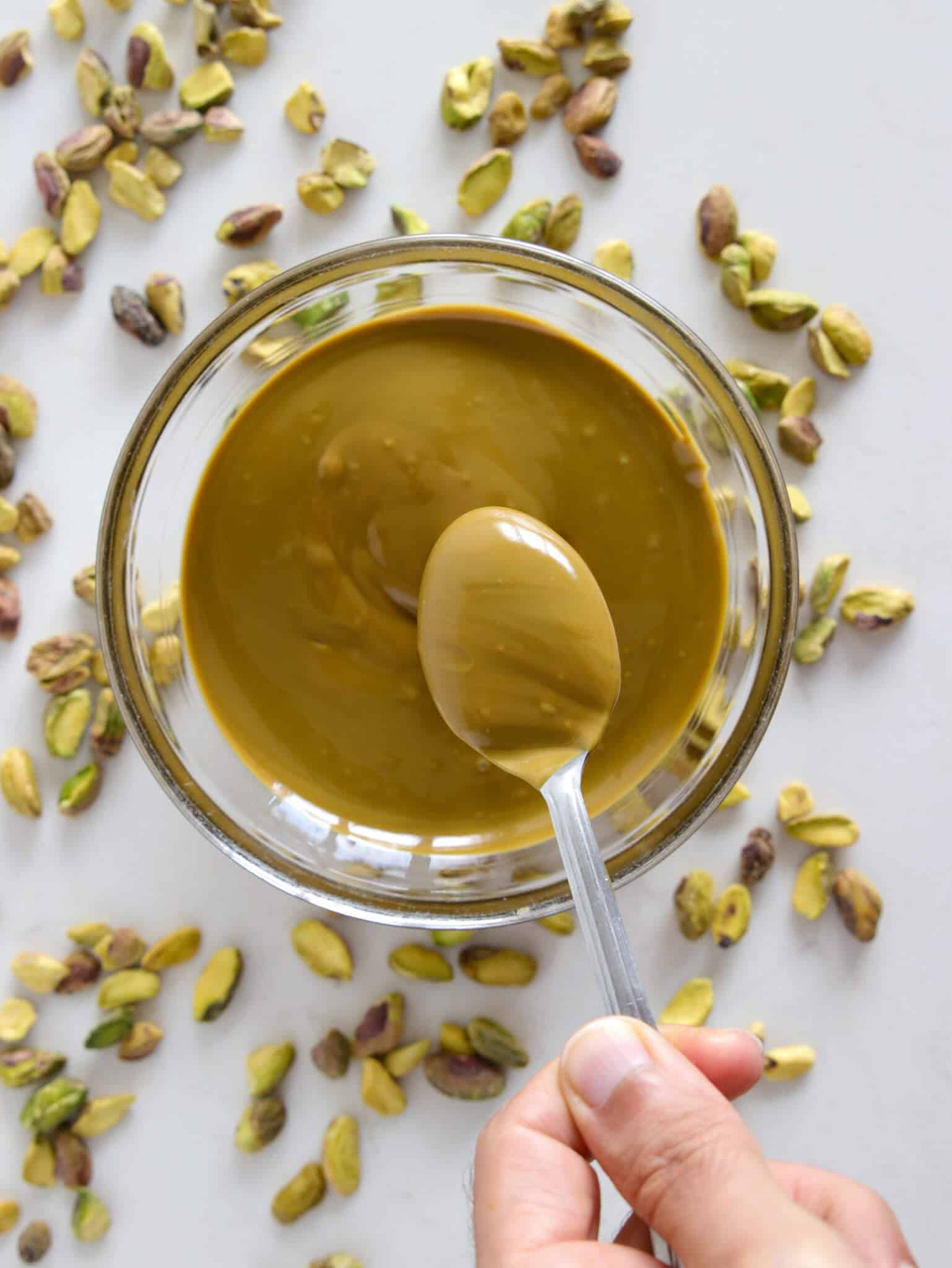 A hand holds a spoon scooping pistachio butter from a glass bowl, surrounded by scattered whole pistachios on a light surface.