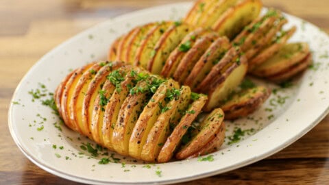 A white plate with sliced, roasted potatoes fanned out and sprinkled with chopped herbs and black pepper, set on a wooden table.