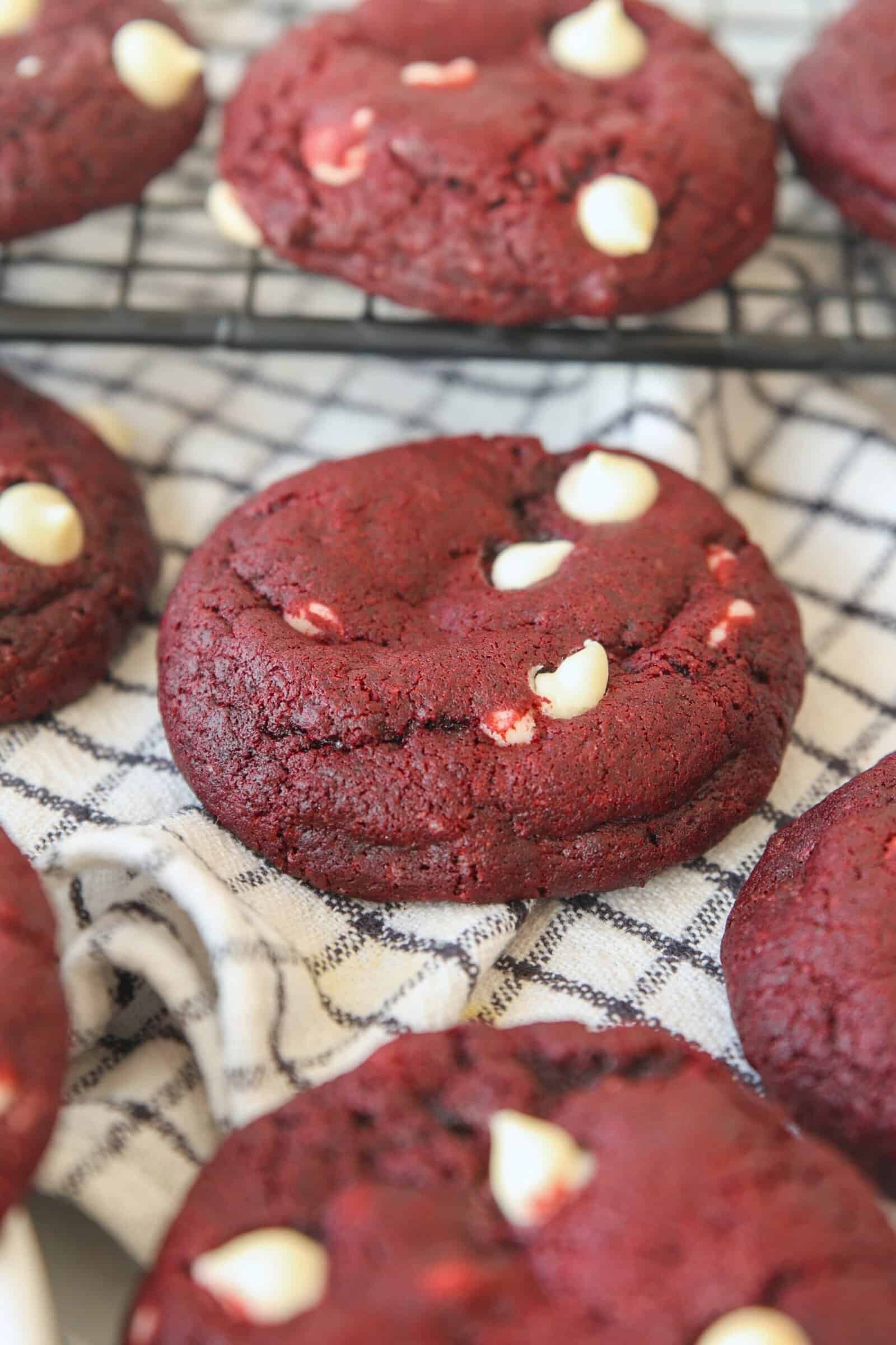 Thick red velvet cookies with white chocolate chips rest on a black and white checkered cloth, with more cookies cooling on a wire rack in the background.