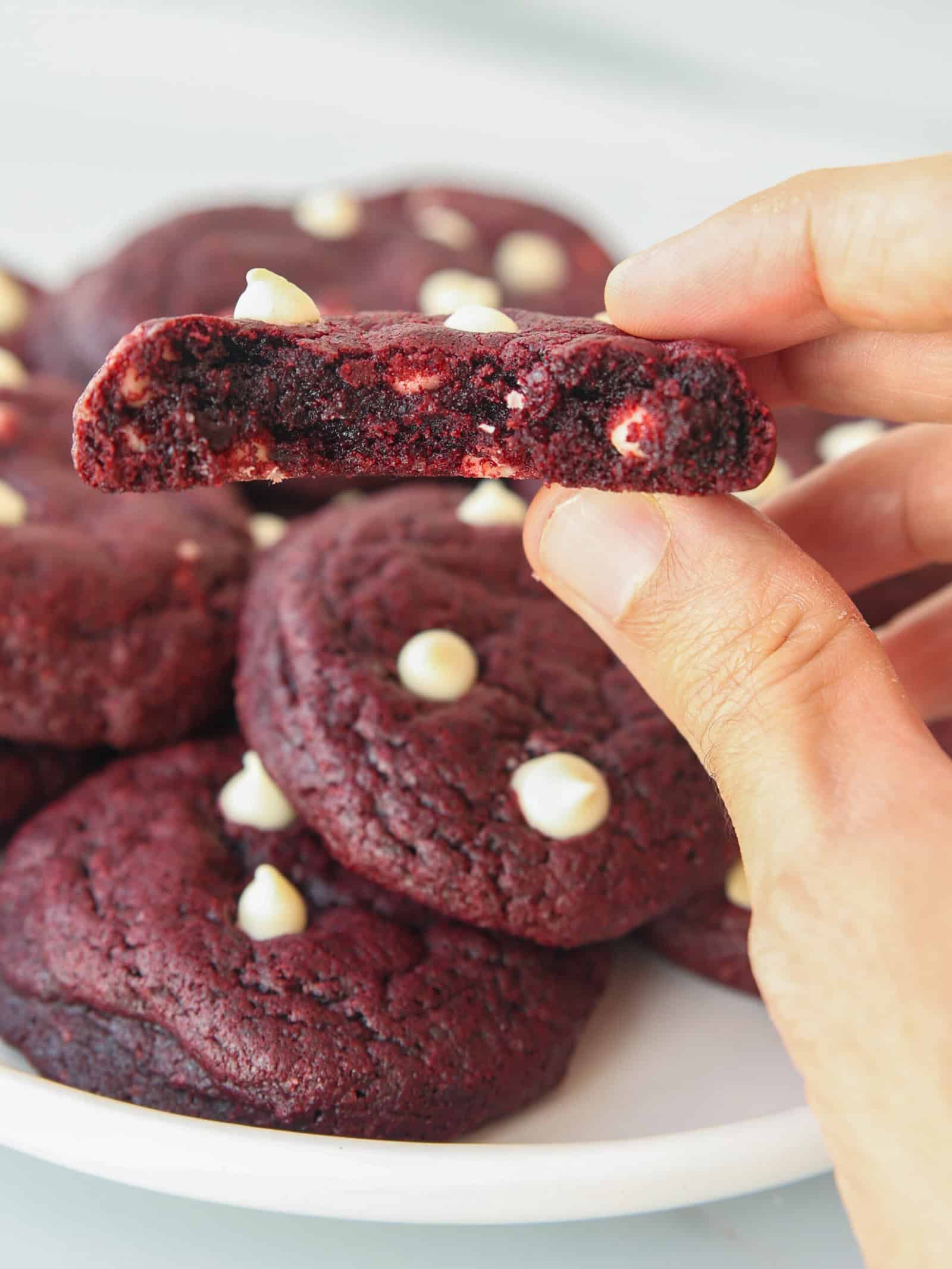 A hand holds a red velvet cookie with white chocolate chips, broken in half to show the soft, gooey center. More cookies with white chocolate chips are stacked on a white plate in the background.
