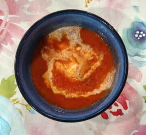 A blue bowl of tomato soup with a swirl of cream on top, placed on a colorful floral-patterned surface.