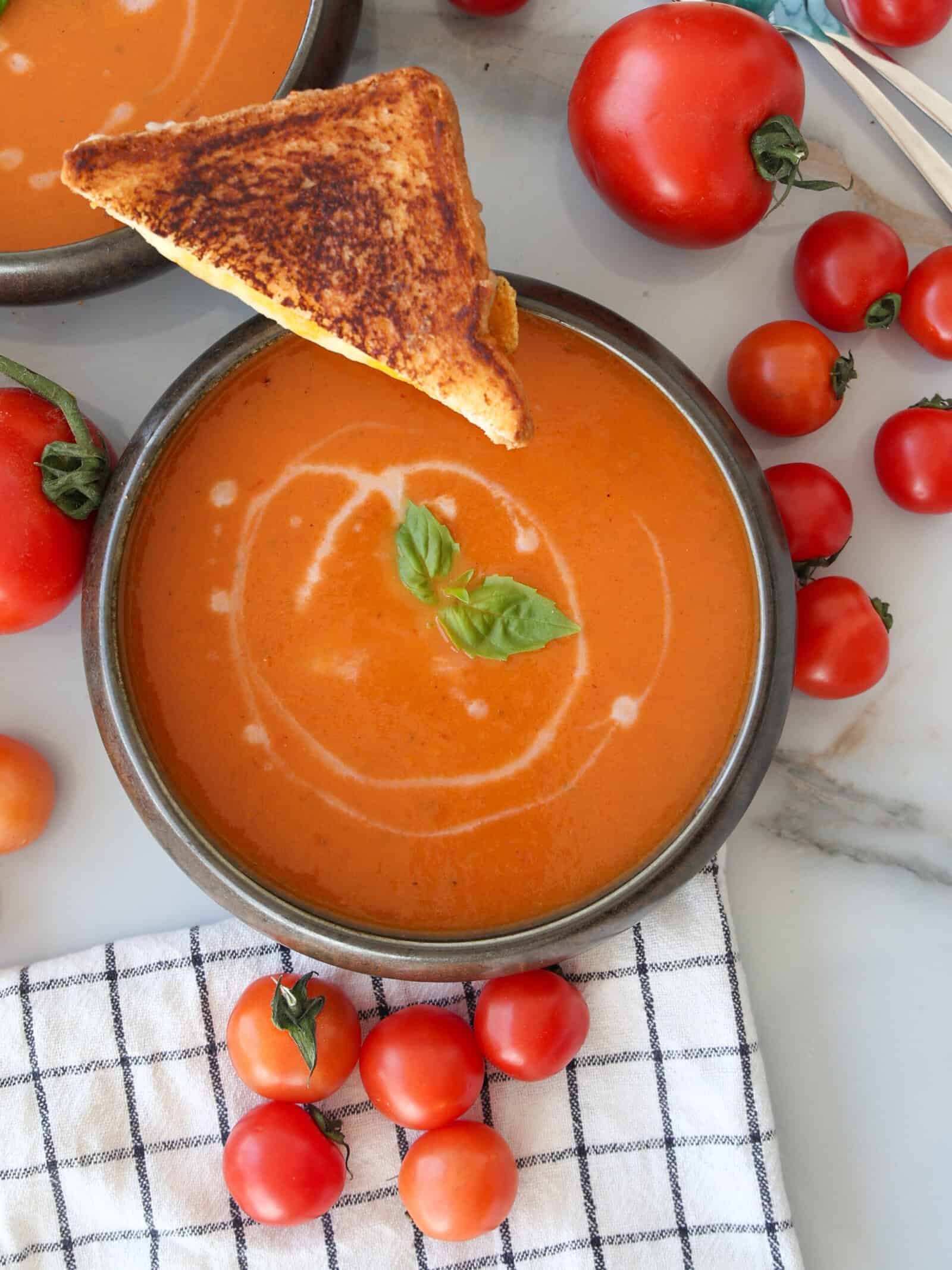 A bowl of creamy roasted tomato soup garnished with basil and a swirl of cream, topped with a triangle of grilled cheese. Fresh tomatoes are scattered around on a checkered cloth and marble surface.