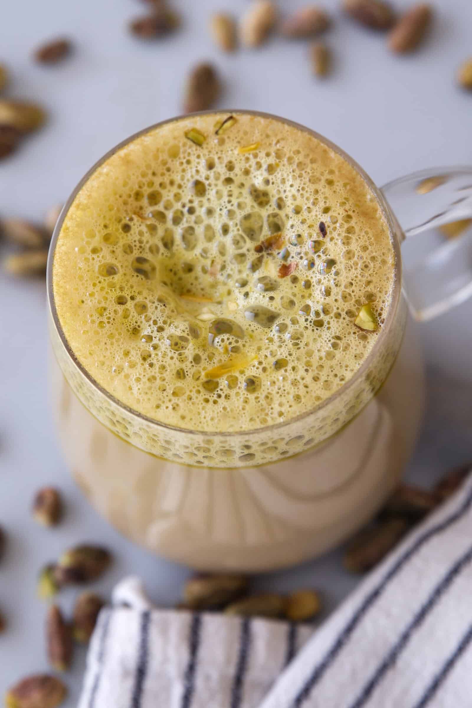 A glass mug filled with frothy pistachio latte, topped with crushed pistachios, sits on a light surface with scattered pistachio nuts and a striped cloth nearby.