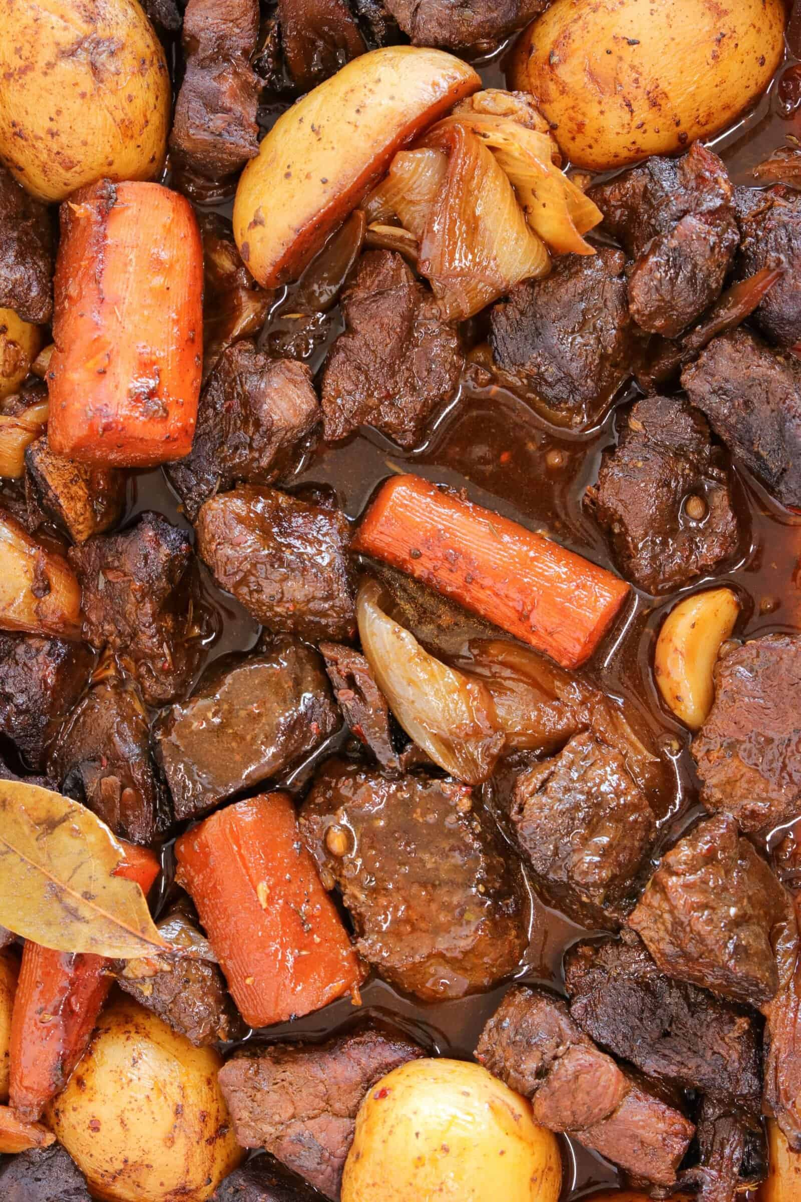 Close-up of a hearty beef stew with chunks of beef, carrots, potatoes, onions, garlic cloves, and bay leaves in a rich, brown sauce.