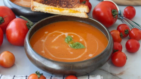 Best Roasted Tomato Soup | Creamy and Easy Recipe A bowl of creamy tomato soup garnished with basil leaves and a swirl of cream, surrounded by fresh tomatoes. A grilled cheese sandwich rests on the edge of the bowl.