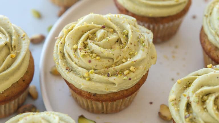 Close-up of cupcakes topped with swirled pistachio frosting and sprinkled with crushed pistachios, displayed on a white plate with some scattered nuts around.