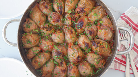 A round metal pot filled with stuffed onions cooked in tomato sauce, garnished with chopped fresh herbs. A red and white striped towel, a fork, and a knife are placed beside the pot.