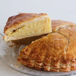 A slice of golden, flaky galette des rois is being lifted from the whole pie, revealing a thick, moist almond filling. The pastry sits on a glass plate with intricate designs.