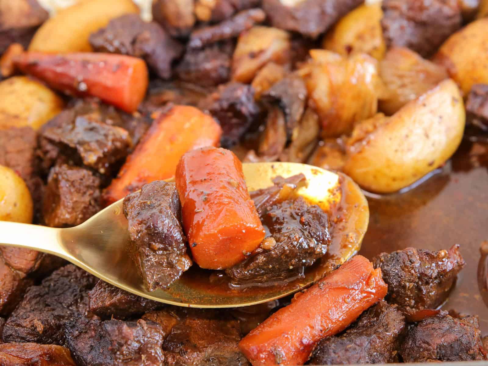 A close-up of a spoon holding chunks of beef, carrots, and a potato from a hearty beef stew with vegetables in a rich brown sauce. More stew ingredients are visible in the background.
