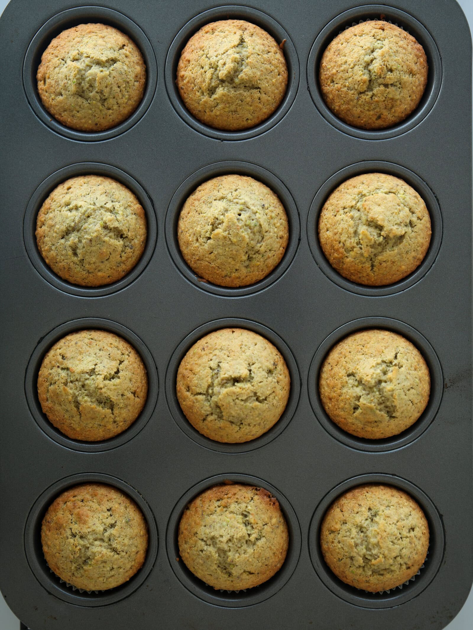 A muffin tin filled with twelve golden-brown muffins, viewed from above. The muffins appear freshly baked and evenly risen in their individual cups.