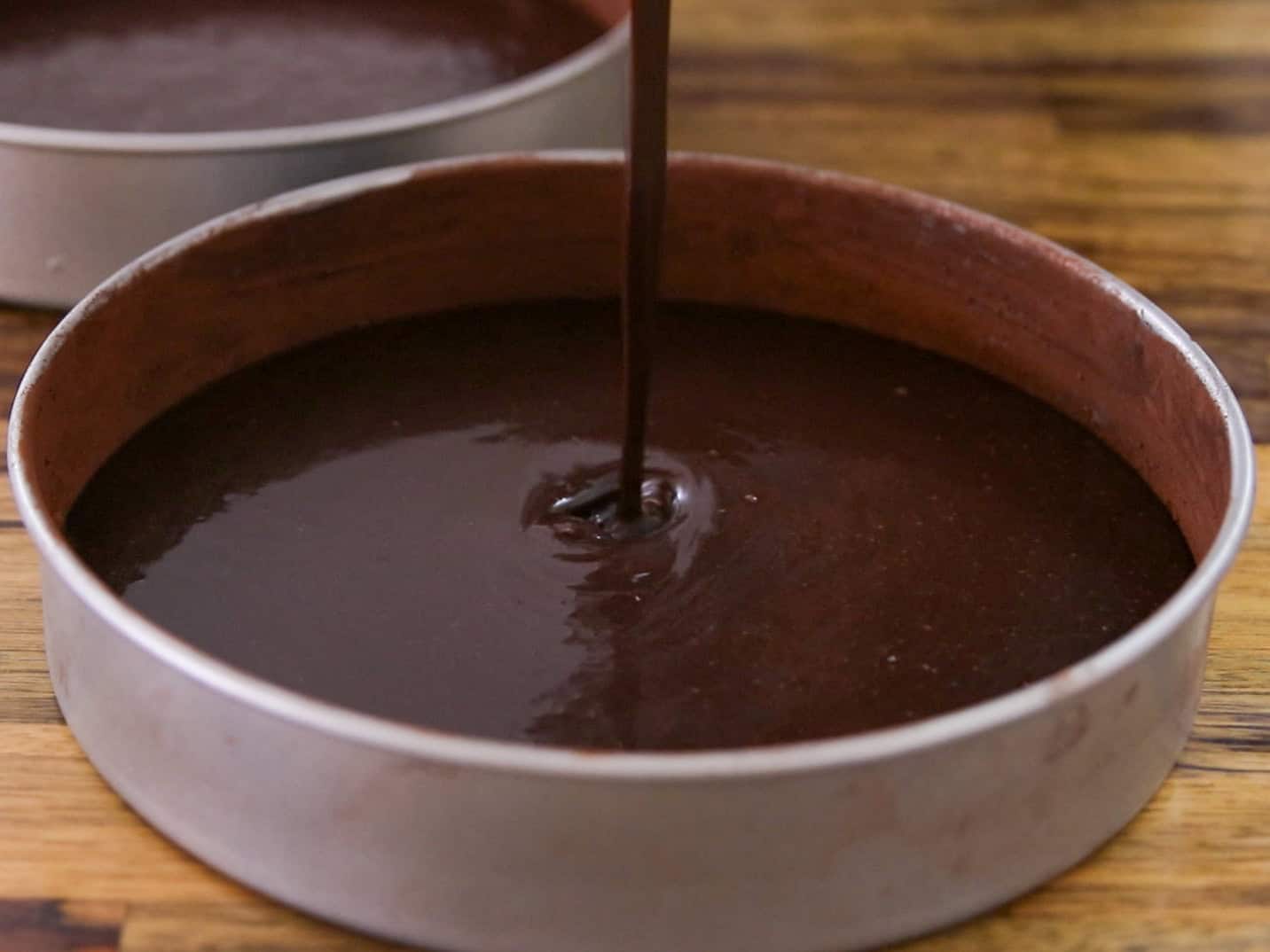 Chocolate cake batter being poured into a round, metal baking pan on a wooden surface. Another pan with batter is visible in the background.