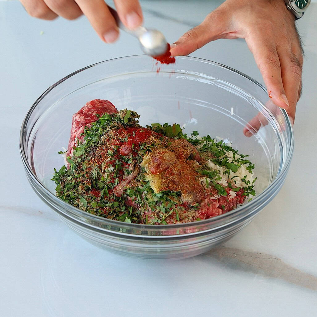 A person sprinkles red spice from a spoon into a glass bowl filled with ground meat, chopped herbs, and seasonings on a white countertop.