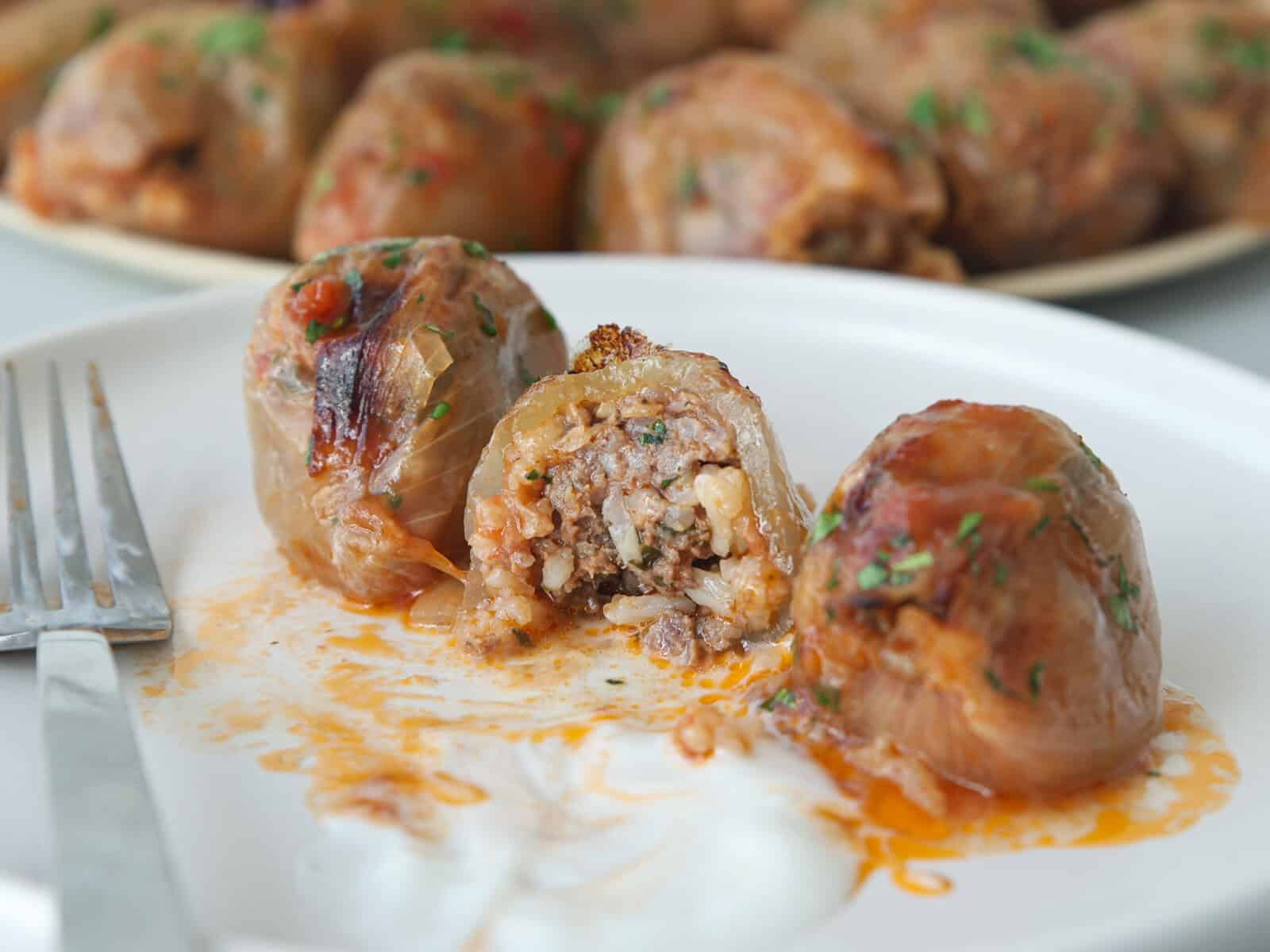 Three onion rolls stuffed with meat and rice on a white plate, one cut open to show the filling. A fork and a dollop of sour cream are on the plate, with more onion rolls blurred in the background.