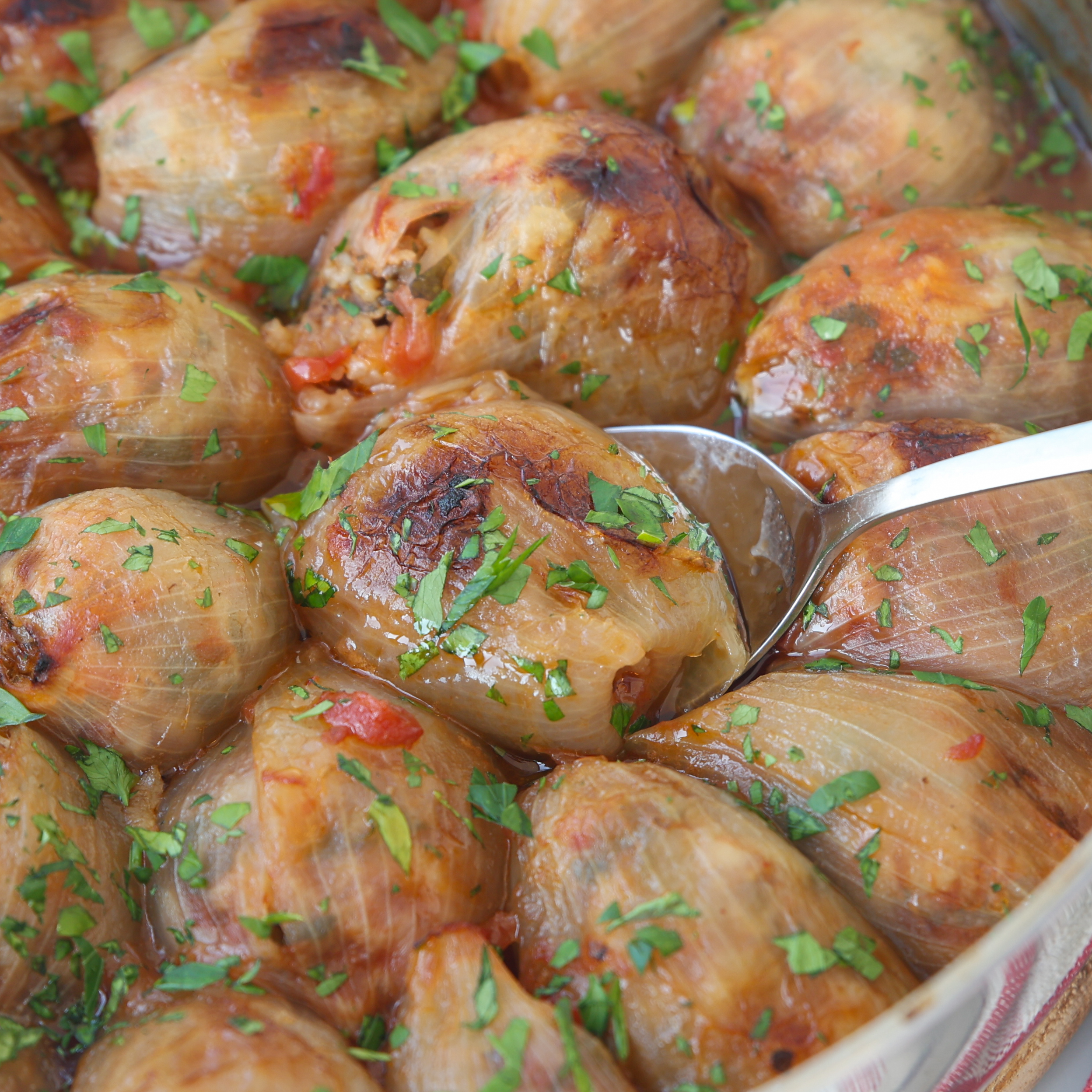 Close-up of stuffed onions baked until golden brown, garnished with chopped parsley. A serving spoon is scooping one of the stuffed onions from the dish.