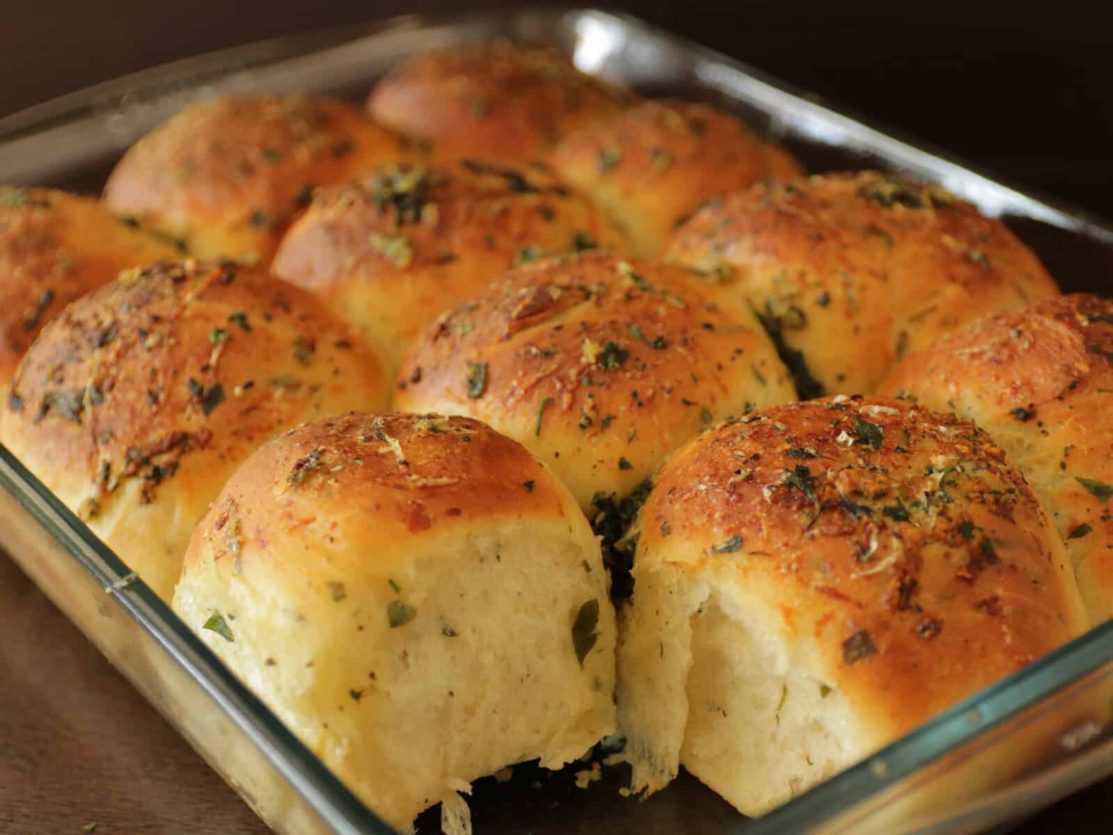 A glass baking dish filled with golden brown, freshly baked garlic dinner rolls topped with herbs. One roll is missing, revealing the soft, fluffy texture inside.