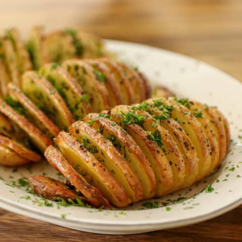 A plate of Hasselback potatoes garnished with chopped herbs, the thinly sliced potatoes are golden brown and seasoned, served on a white dish against a wooden background.