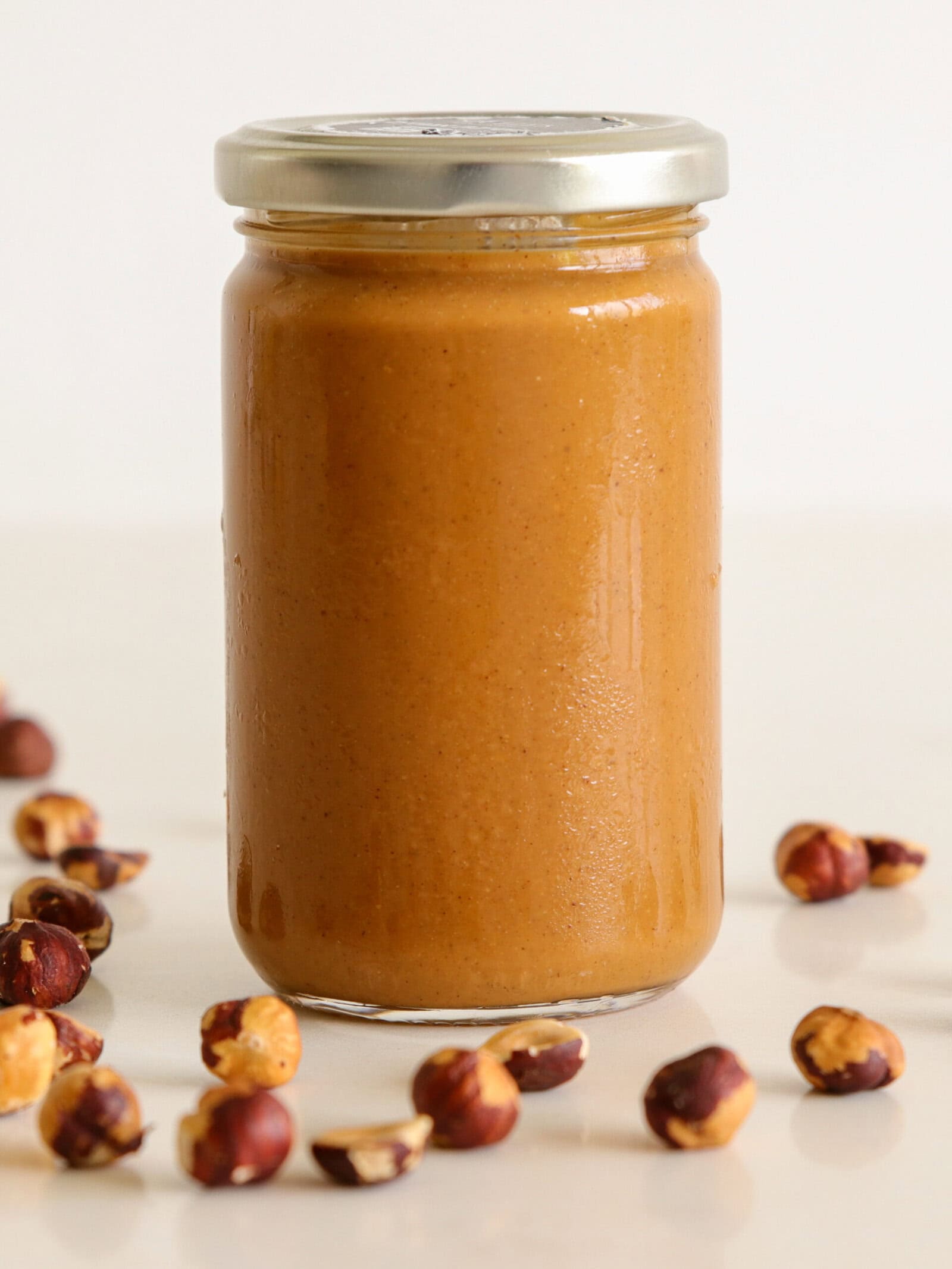 A glass jar filled with creamy brown nut butter, sealed with a metal lid, surrounded by scattered whole hazelnuts on a white surface.