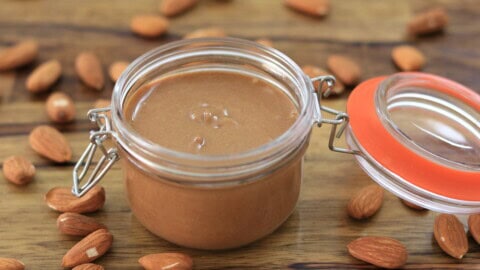 A glass jar filled with creamy almond butter sits open on a wooden surface, surrounded by scattered whole almonds.