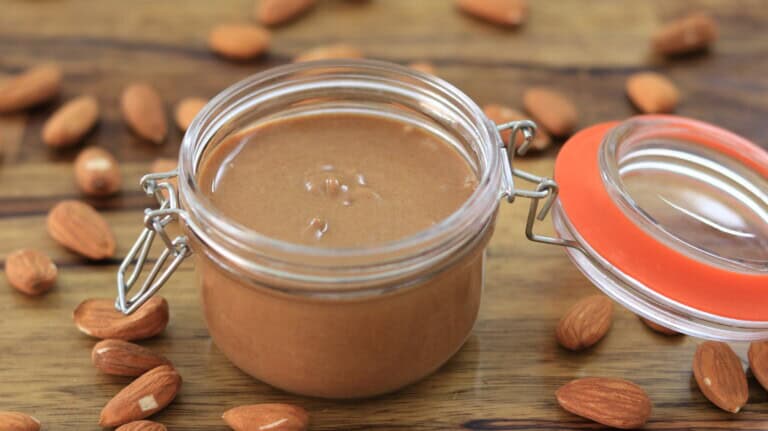 A glass jar filled with creamy almond butter sits open on a wooden surface, surrounded by scattered whole almonds.