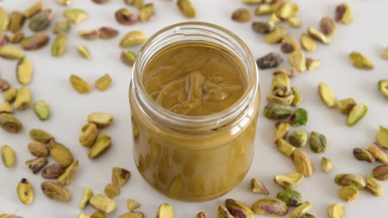 A glass jar filled with smooth pistachio butter sits on a white surface, surrounded by scattered whole and shelled pistachios.