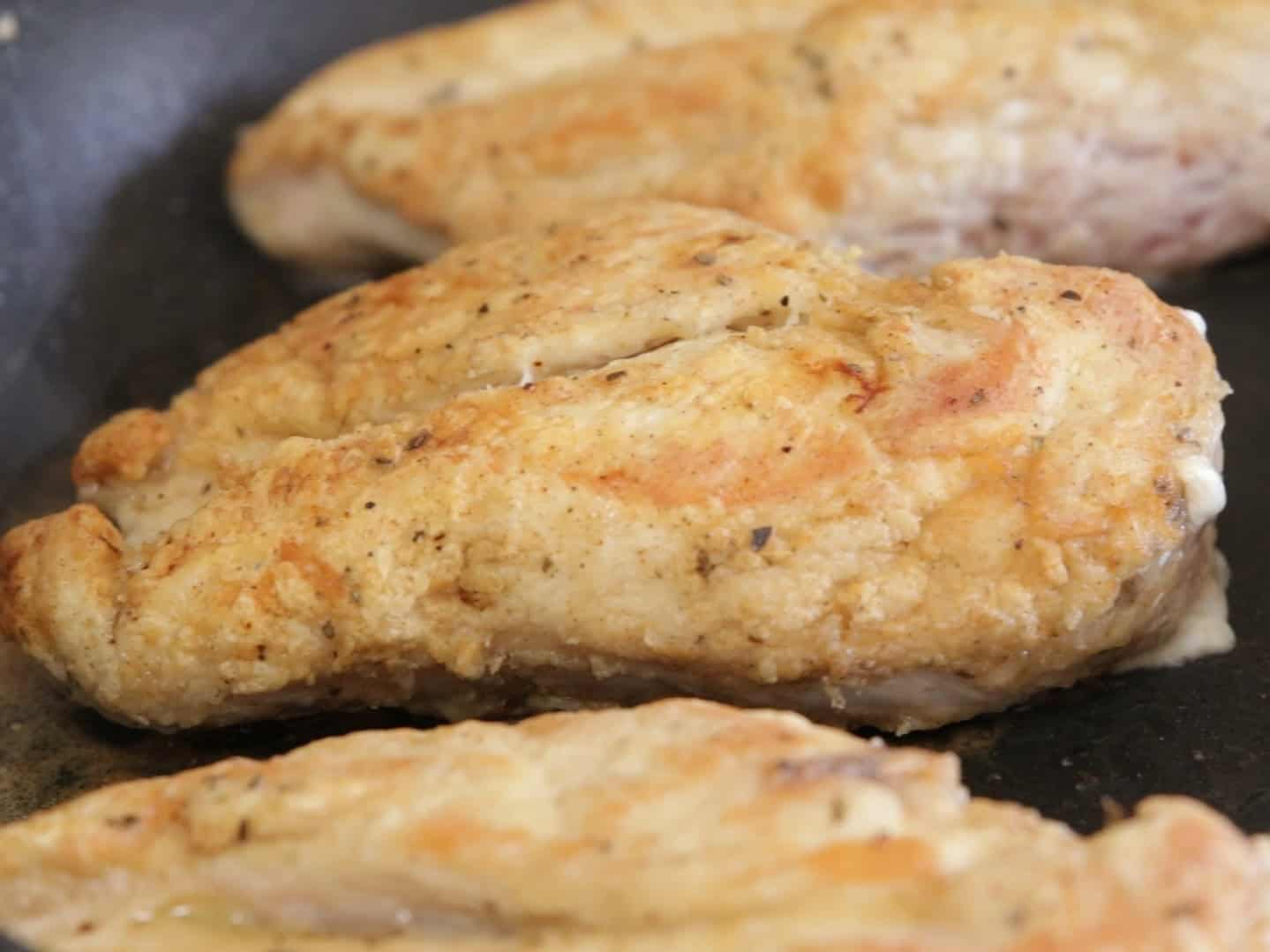 Close-up of three seasoned and cooked chicken breasts in a pan, with a golden-brown crust visible on the surface.