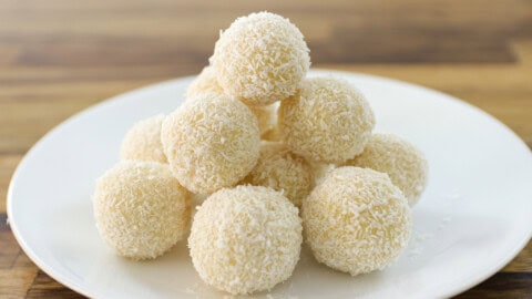 Easy White Chocolate Truffles | Rich and Creamy A white plate holds a pyramid of round, white coconut-covered sweets, stacked neatly. The background features a wooden surface.