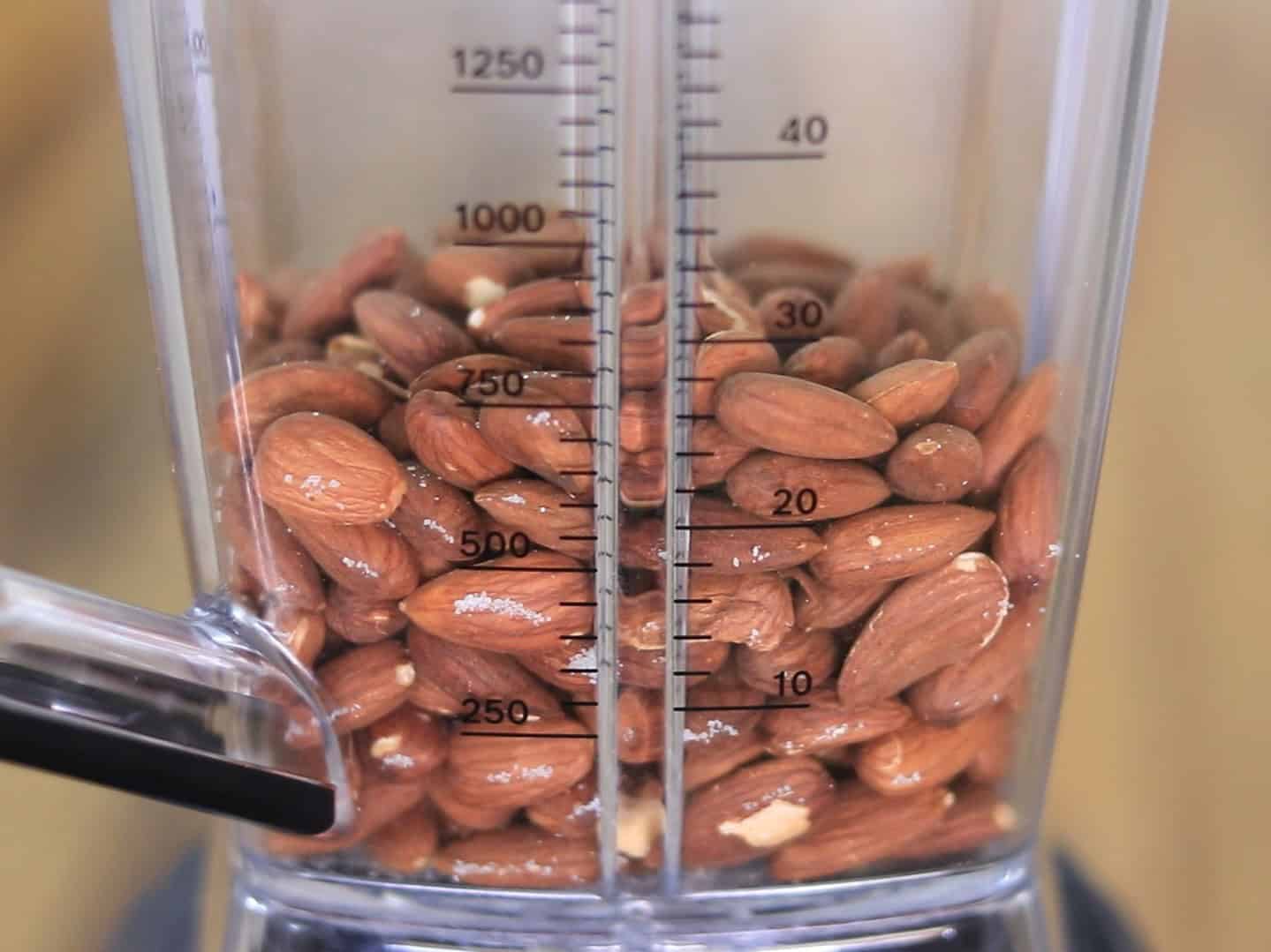 Whole almonds inside a blender jar, with measurement markings visible on the container. The almonds are piled up to around the 300 ml mark.