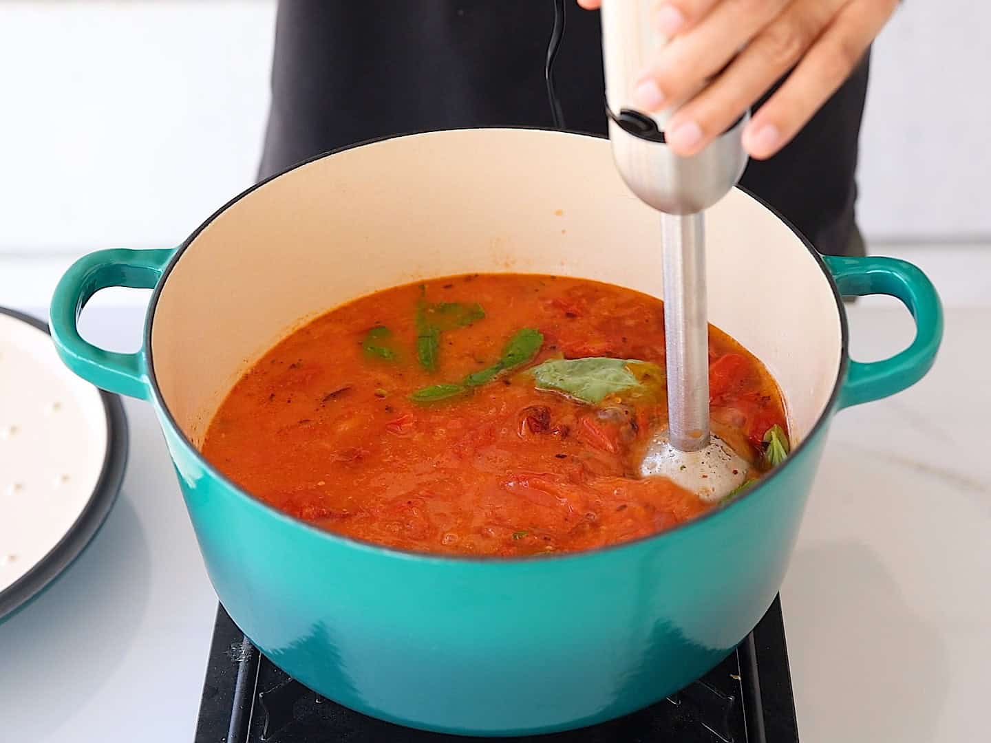 A person uses an immersion blender to blend tomato soup with basil leaves in a large turquoise pot on a stovetop.