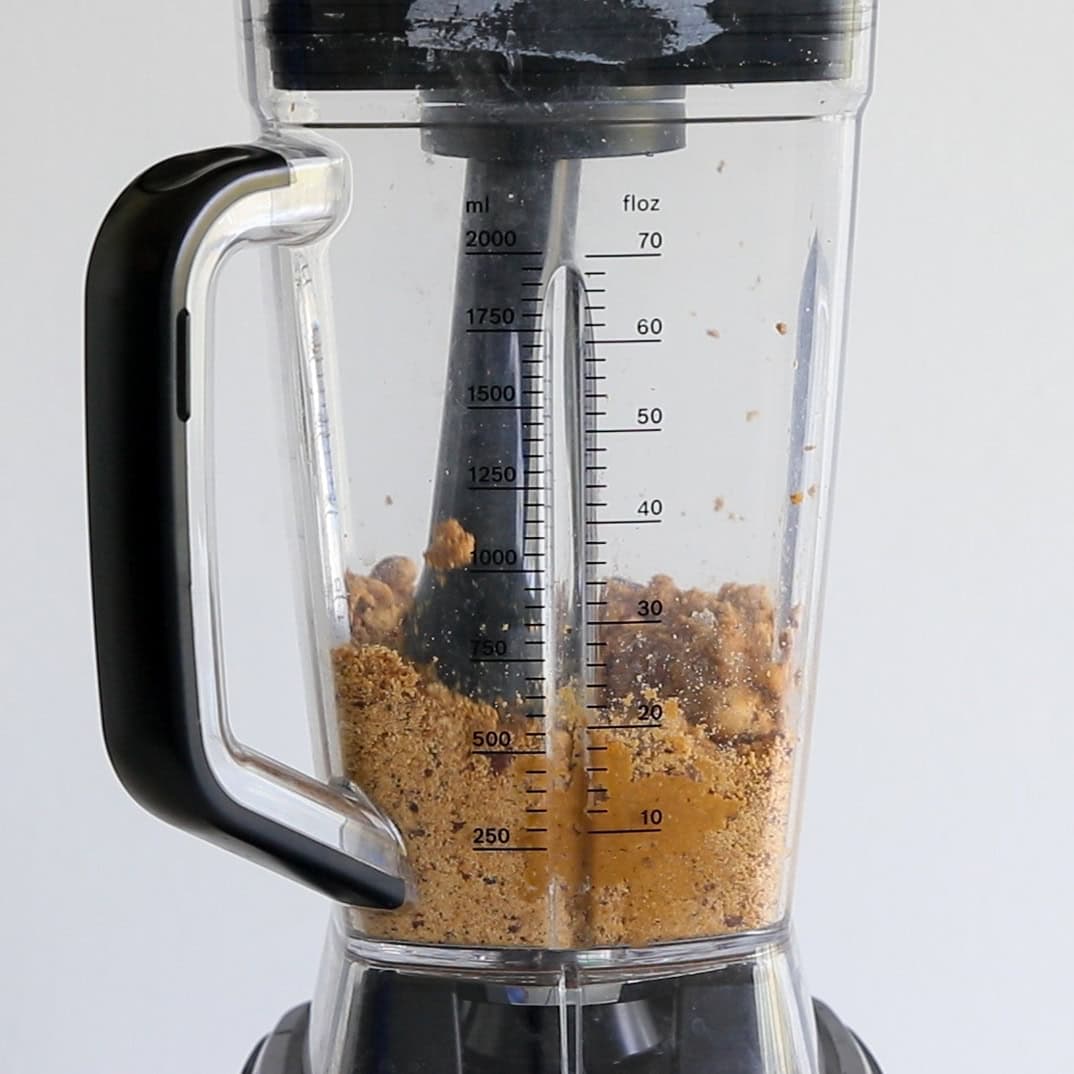 A close-up of a blender containing crushed cookies or biscuits, seen through the transparent container marked with measurements in milliliters and fluid ounces. The blender is placed against a plain white background.