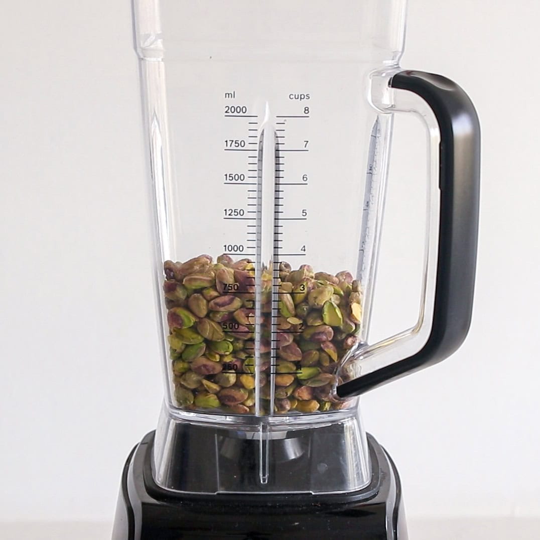 A blender filled with unshelled pistachios, positioned on a countertop against a plain white background. The blender jar shows measurement markings in milliliters and cups.