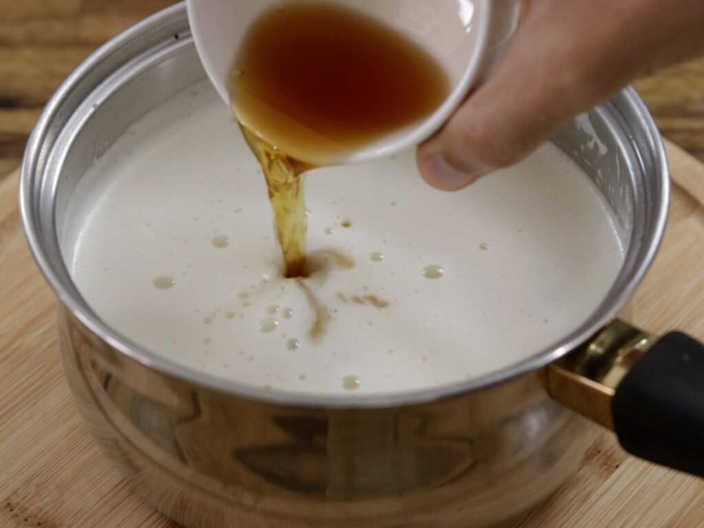 A hand pours a small cup of brown liquid into a saucepan filled with a creamy white mixture, set on a wooden surface.
