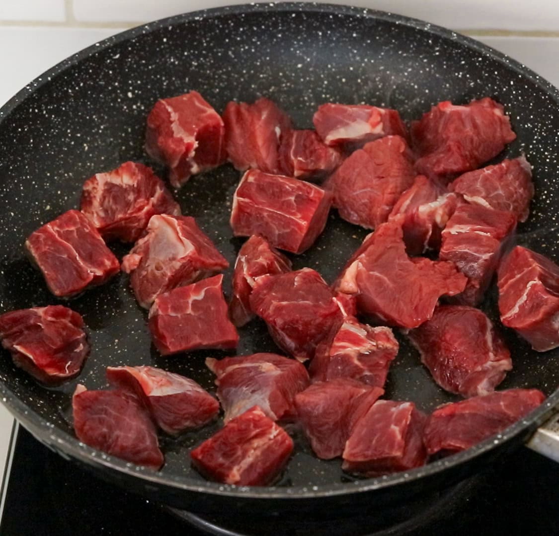 Chunks of raw beef are spread out in a black, nonstick frying pan, ready to be cooked. The pan is placed on an electric stovetop.