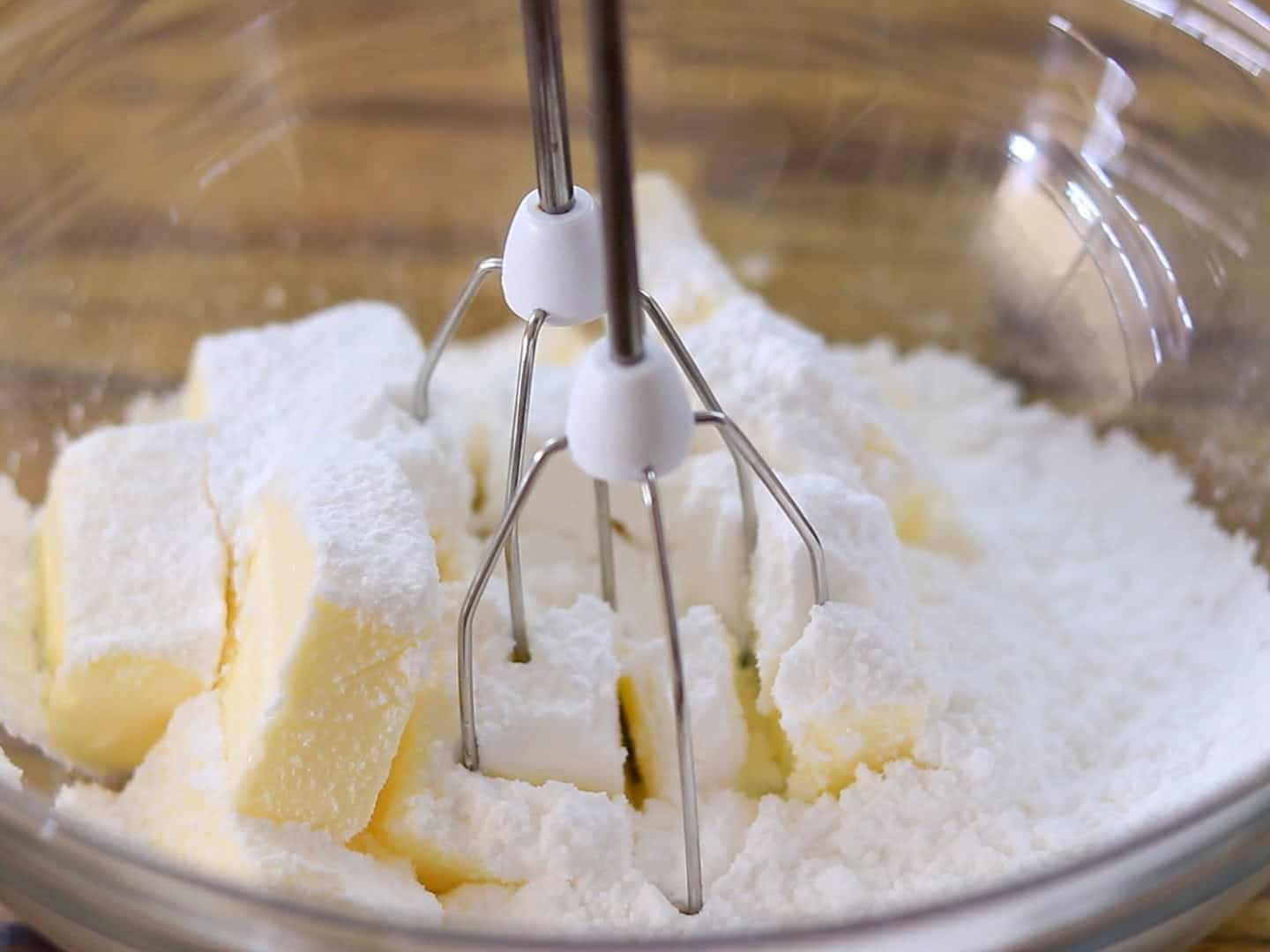 A close-up of an electric hand mixer in a glass bowl with cubed butter and powdered sugar, ready to be mixed.