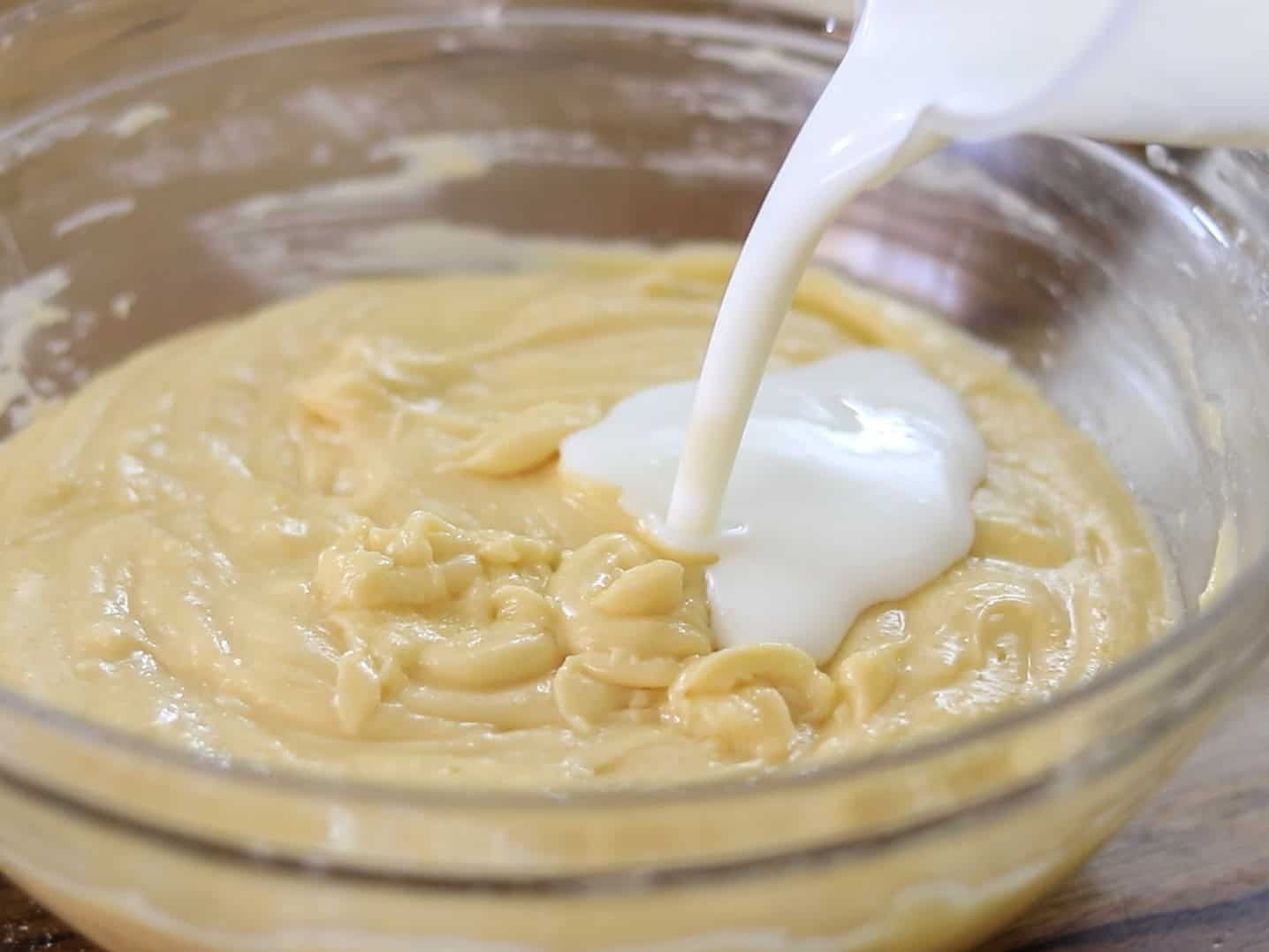 A close-up of buttermilk being poured into a glass bowl filled with yellow cake batter, ready to be mixed.