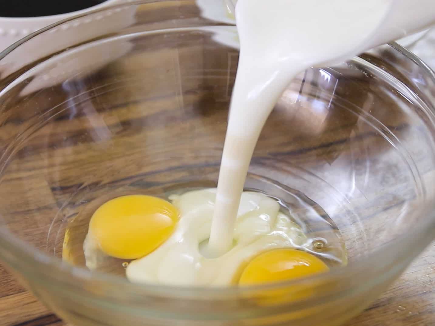 A glass bowl with two raw eggs is shown as buttermilk or cream is being poured in, ready for mixing. The bowl is on a wooden surface.