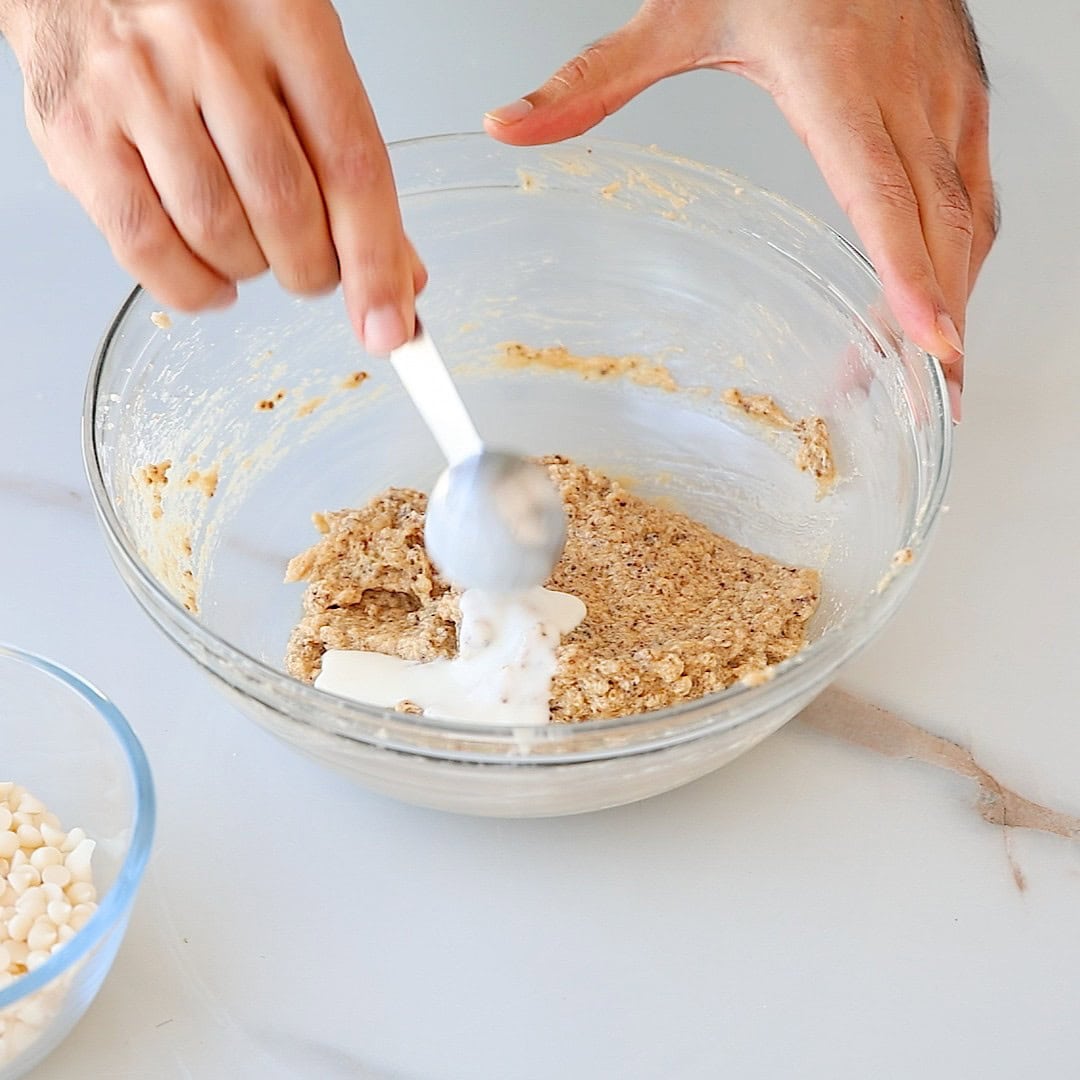 A person mixes a thick batter in a glass bowl with a spoon, while a small bowl of white chocolate chips sits nearby on a white countertop.