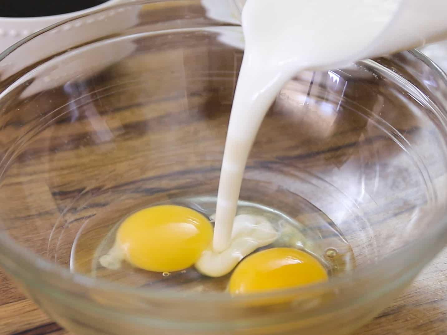 A glass bowl with two raw eggs inside,  buttermilk is being poured in from a measuring cup, all placed on a wooden surface.