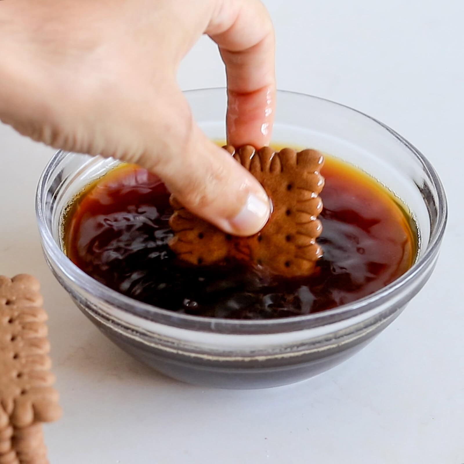 A hand dips a rectangular chocolate biscuit into a glass bowl filled with dark coffee on a white surface.