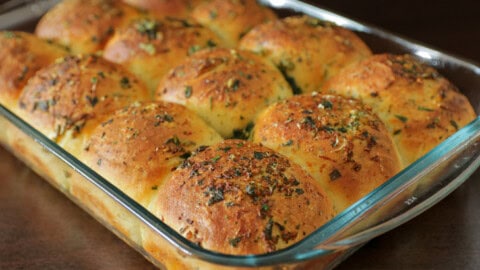 A glass baking dish filled with golden-brown, freshly baked dinner rolls topped with herbs and seasonings, arranged closely together on a dark surface.