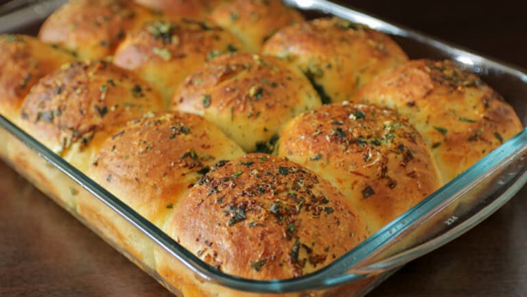 A glass baking dish filled with golden-brown, freshly baked dinner rolls topped with herbs and seasonings, arranged closely together on a dark surface.