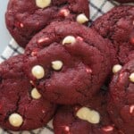 A close-up of several red velvet cookies with white chocolate chips, stacked on a striped cloth. The cookies have a rich red color and soft, chewy texture.