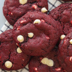 A close-up of several red velvet cookies with white chocolate chips, stacked on a striped cloth. The cookies have a rich red color and soft, chewy texture.