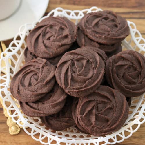 A white decorative plate holds several round, swirled chocolate cookies arranged neatly. A gold fork and a white teacup sit nearby on a wooden surface.