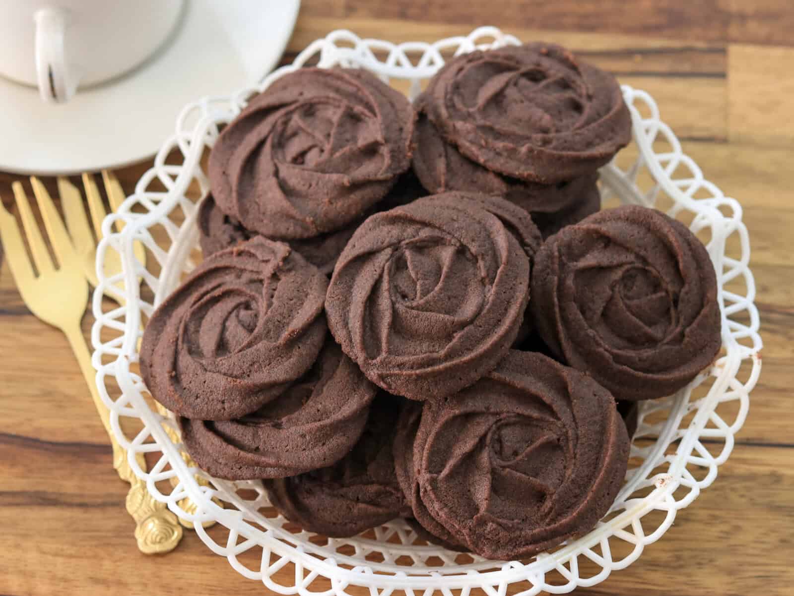 A white decorative plate holds several round, piped chocolate butter cookies arranged neatly. A gold fork and a white teacup sit nearby on a wooden surface.