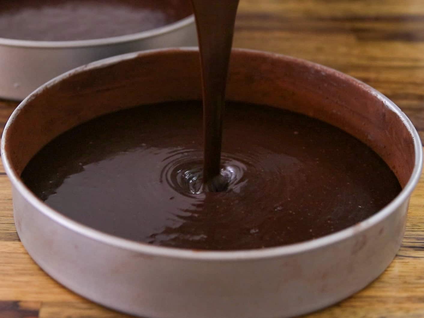 Chocolate cake batter being poured into a round metal baking pan on a wooden surface.
