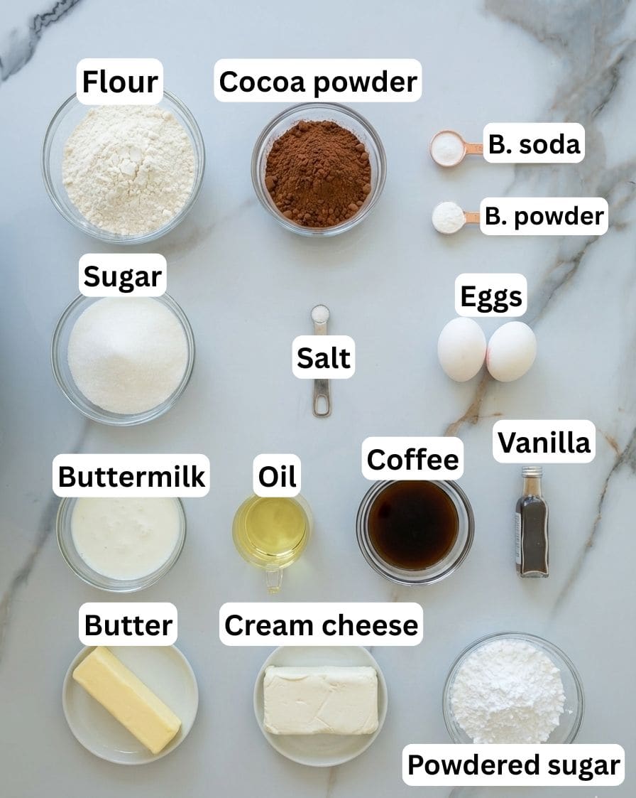 Labeled ingredients for baking, including flour, cocoa powder, baking soda, baking powder, sugar, eggs, salt, buttermilk, oil, coffee, vanilla, butter, cream cheese, and powdered sugar on a light surface.